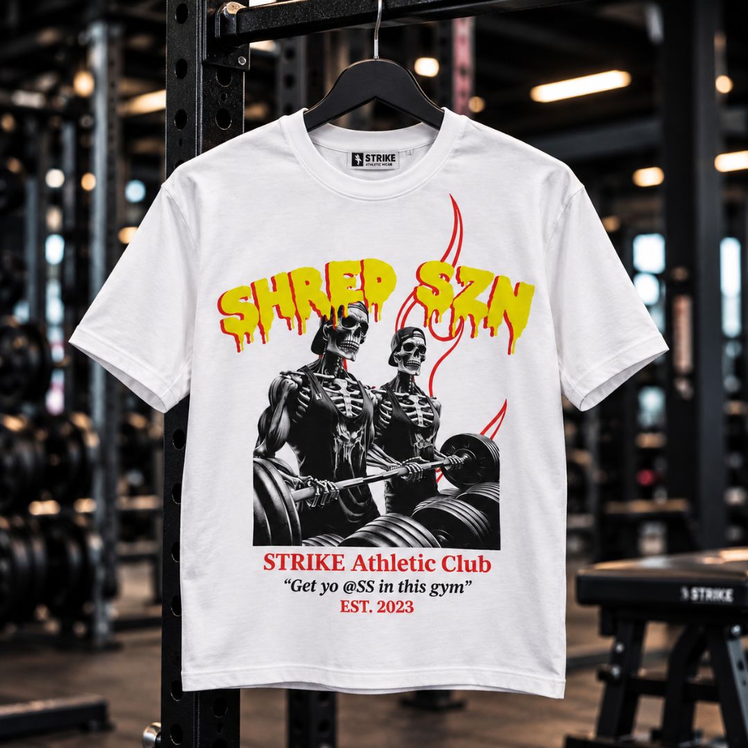 A white graphic t-shirt hanging on a black rack inside a gym. The shirt features a black and white graphic of two skeleton gym rats lifting weights, with yellow dripping text reading 'SHRED SZN' above and red text below reading 'STRIKE Athletic Club' and a slogan.