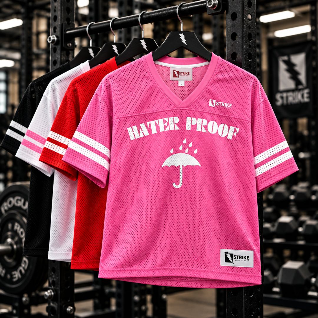 Three mesh V-neck athletic jerseys hang on a rack in a gym: a bright pink one in front with white 'HATER PROOF' text and umbrella graphic, plus white sleeve stripes; a red one behind it with white sleeve stripes; and a black-and-white one at the back with pink sleeve stripes. The jerseys show the Strike brand logo.