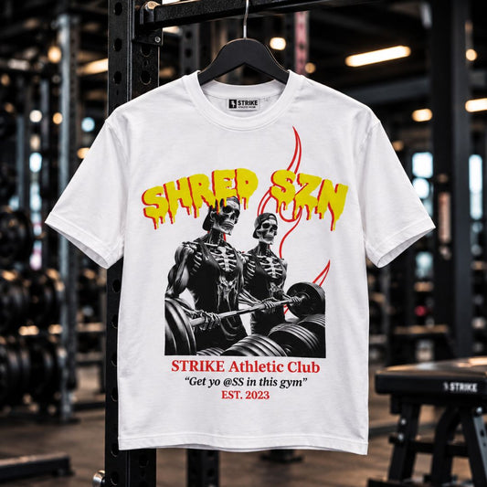 A white graphic t-shirt hanging on a black rack inside a gym. The shirt features a black and white graphic of two skeleton gym rats lifting weights, with yellow dripping text reading 'SHRED SZN' above and red text below reading 'STRIKE Athletic Club' and a slogan.