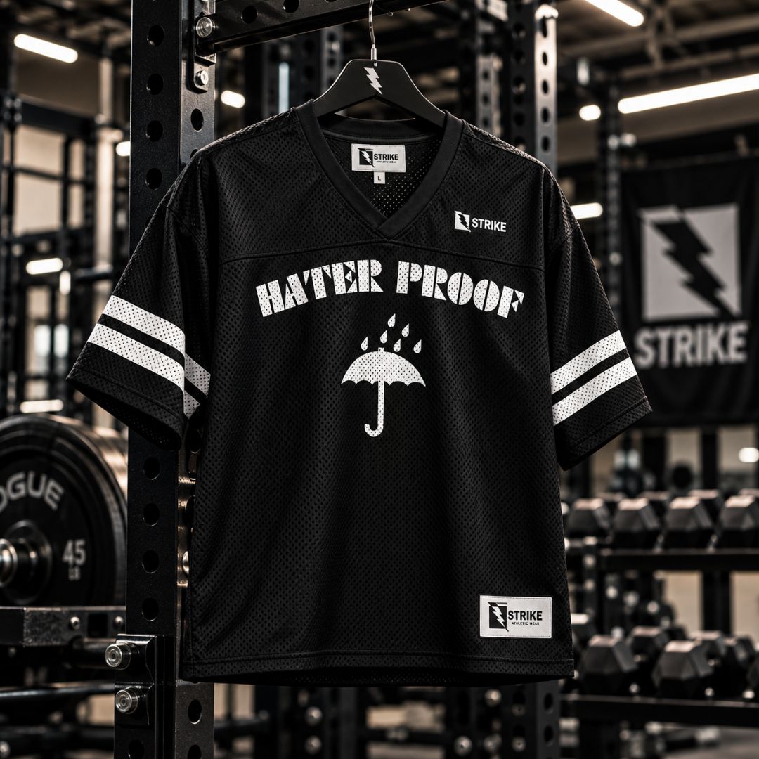 A black mesh V-neck athletic jersey with white sleeve stripes and a white 'HATER PROOF' graphic with an umbrella design, hanging on a rack in a gym.