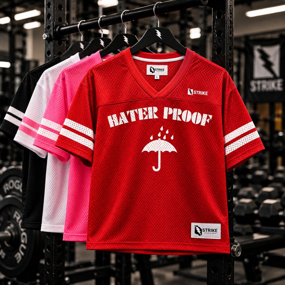 A red mesh V-neck jersey shirt hanging on a rack in a gym, with white sleeve stripes and a white 'HATER PROOF' graphic featuring an umbrella. Other color variants in black, white, and pink are visible behind it.